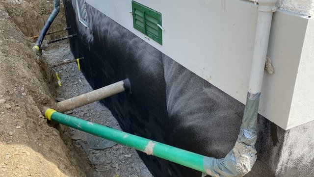 Freigelegte Kellerwand mit schwarzer Masse und Drainagerohren im Erdgraben