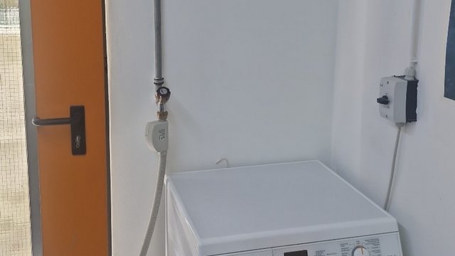 Weiße Waschmaschine steht in Raumecke neben oranger Tür, Zulaufschlauch führt an weißer Wand herab.