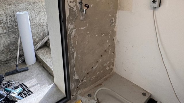 Graue Wand mit Bohrlöchern und abgeplatzter Masse, davor Werkzeug am Boden