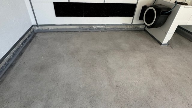 Grauer Estrichboden mit Randabdichtung und schwarzer Paneelwand im Eckbereich
