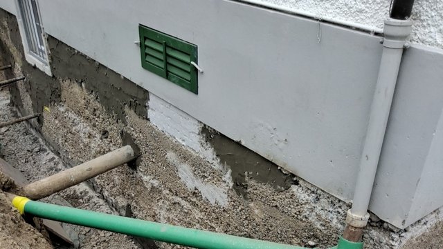 Freigelegte Gebäudesockelwand mit rauer grauer Masse und grünem Lüftungsgitter