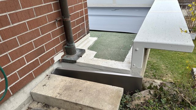 Metallabdeckung an Lichtschacht neben Ziegelmauer und Fallrohr, Betonstufe vorn.