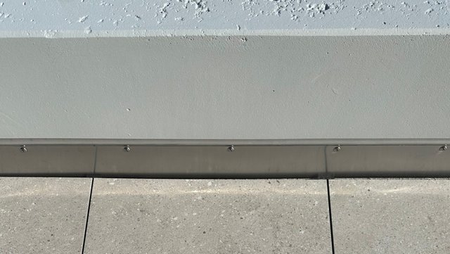 Verschraubte Metallleiste am Wandfuß über grauen Bodenplatten, Nahaufnahme