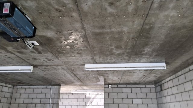 Beton­decke mit Bohrlöchern, Feuchtflecken und zwei Leuchtstofflampen in Raum aus Betonsteinen
