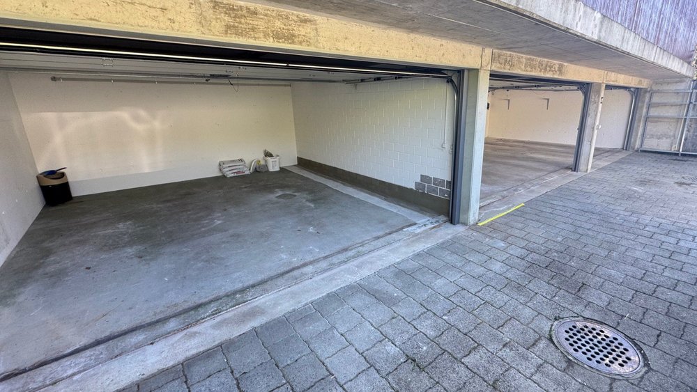 Offene Garagenstellplaetze mit Betonboden, weisse Waende und Pflasterflaeche vorn.