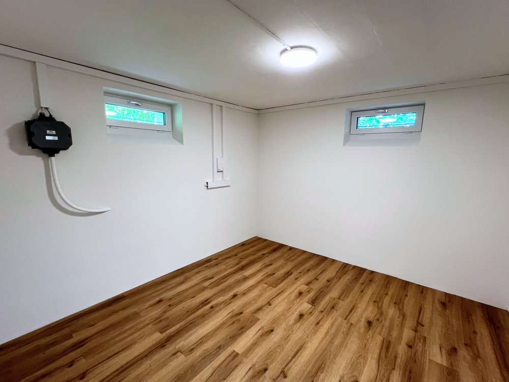 Leerer Kellerraum mit zwei kleinen Fenstern, weißer Decke und Holzoptikboden.