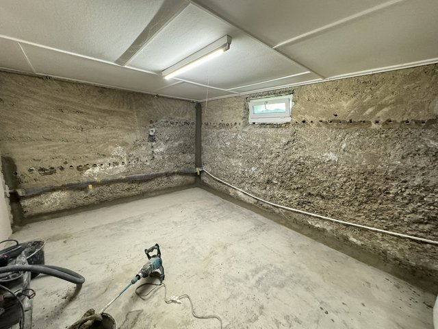 Kellerraum mit freigelegter Betonwand, Bohrlochreihe und Bohrhammer auf Betonboden