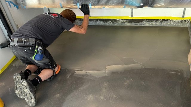 Arbeiter kniet am Boden und glättet graue Masse entlang einer Kante.