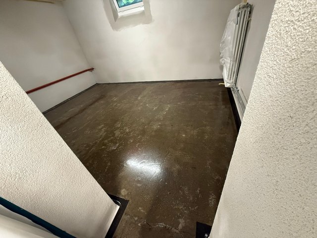 Leerer Kellerraum mit dunklem Betonboden, Heizkörper und kleinem Fenster