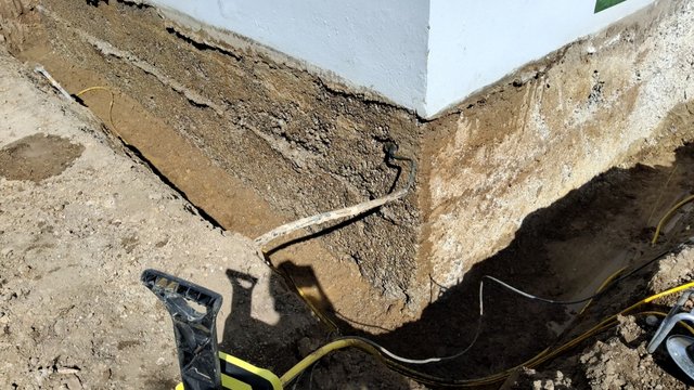 Freigelegte Baugrube entlang Hauswand mit Erdreich, Kabeln und Spaten