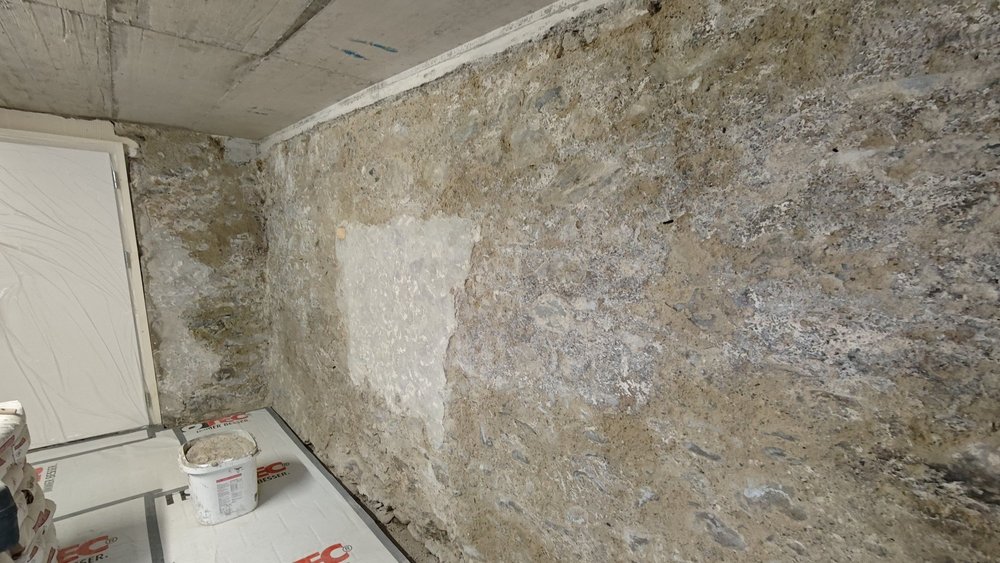 Kellerwand aus grobem Mauerwerk mit hellem Reparaturfleck, Boden mit Abdeckplatten und Eimer