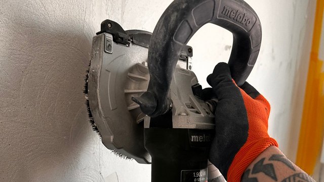 Hand führt Schleifmaschine mit Bürstenkranz an rauer Wand entlang
