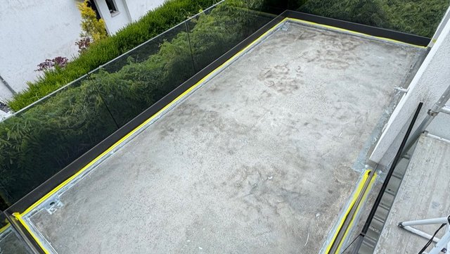 Freigelegte Balkonfläche mit grauer Betonoberfläche und gelbem Randband, Glasgeländer