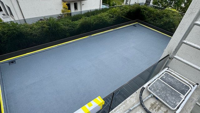 Graue Abdichtungsbahn auf Flachdach mit gelb markiertem Rand, Blick von Gerüst