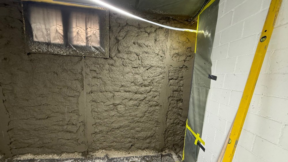 Graue Spritzmasse an Kellerwand unter kleinem Fenster, daneben weiße Mauersteine und Wasserwaage