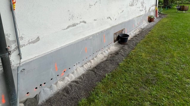 Bohrlöcherreihe und graue Masse am Sockel eines Gebäudes, daneben Kiesstreifen und Rasen.