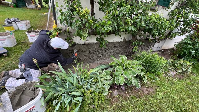 Arbeiter trägt graue Masse auf Sockelbereich einer Hauswand im Garten auf.