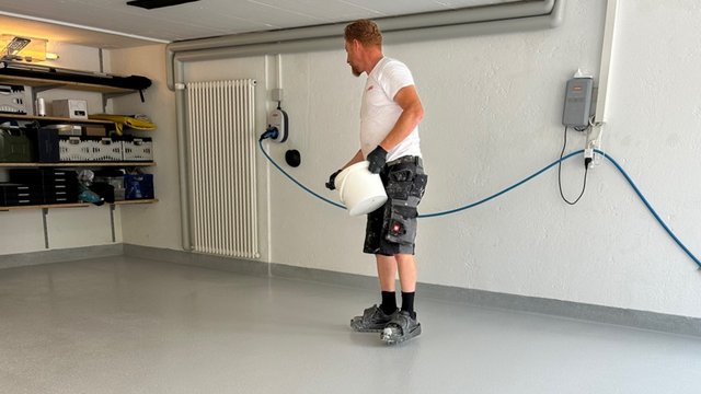 Arbeiter verteilt transparente Flüssigkeit auf grauem Boden in einem Raum.