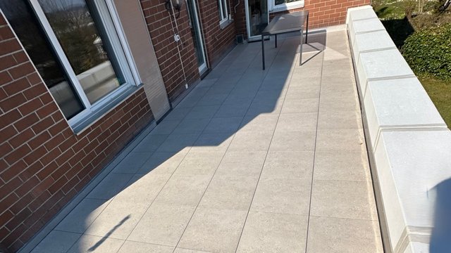 Gefliester Balkon mit Ziegelwand, Fenstern, Tisch und weißer Brüstung bei Sonne