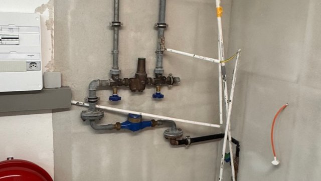 Druckminderer und blauer Wasserzähler an grauen Stahlrohren vor grauer Wand