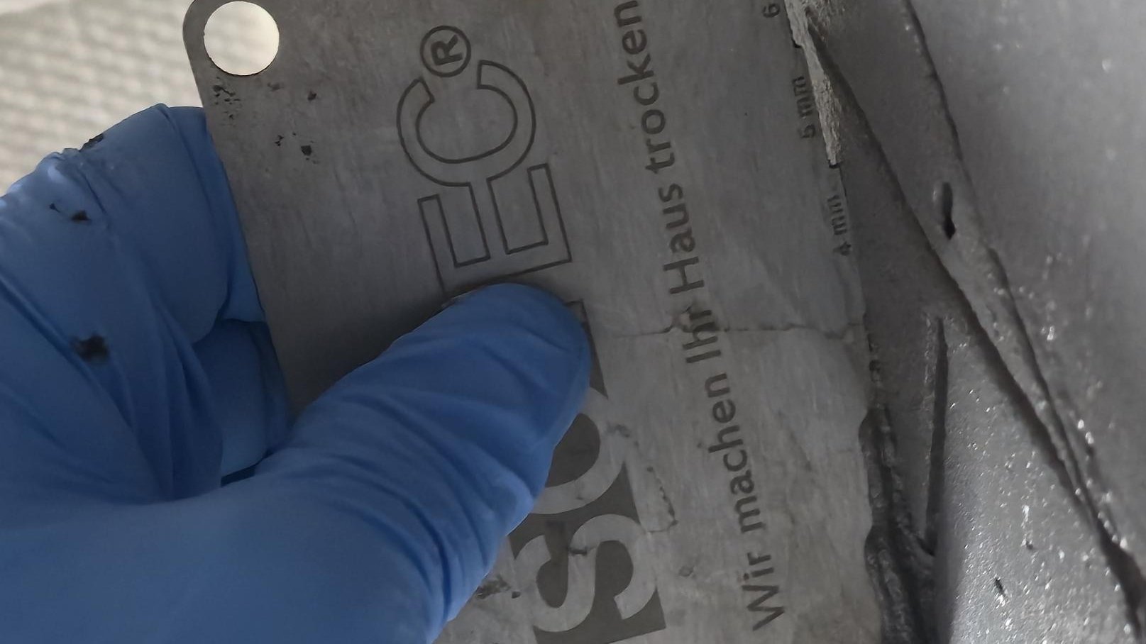 Blaue Handschuhe halten EC‑Karte und ziehen schwarze Masse entlang einer Fuge.
