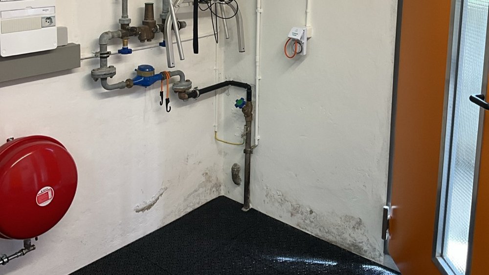 Technikraum mit weißer Wand, Rohrleitungen, blauem Wasserzähler und rotem Ausdehnungsgefäß