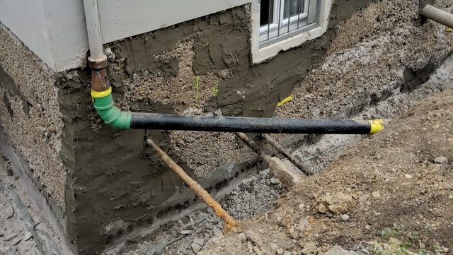 Freigelegte Gebäudewand mit grauer Masse, Drainrohr und Kiesbett