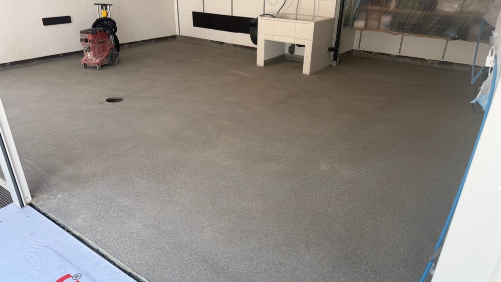 Frisch geglätteter grauer Boden mit Abflussöffnung, Baustellengerät im Hintergrund