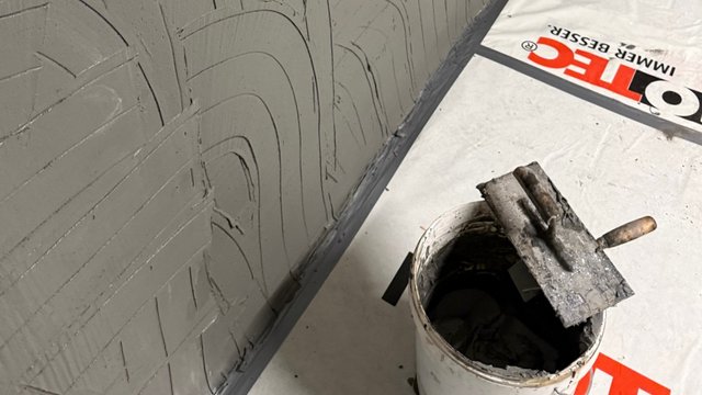 Maurerkelle mit grauer Masse auf Eimer neben Wand mit grauer Spachtelstruktur
