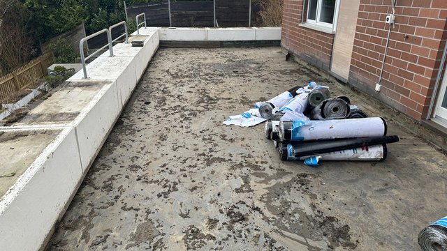Dachterrasse mit verschmutzter grauer Fläche und gerollten schwarzen Bahnen neben Ziegelwand