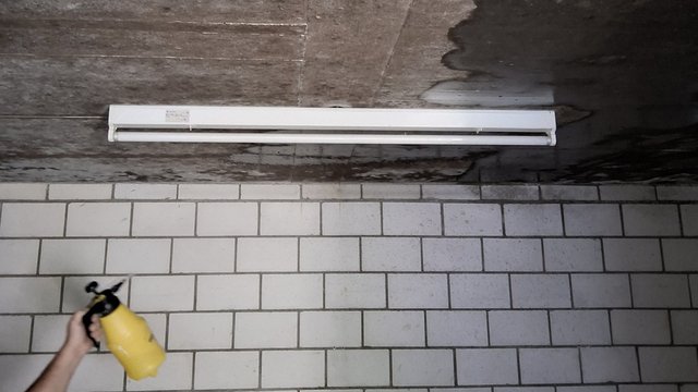 Hand sprüht transparente Flüssigkeit auf verfärbte Betondecke über gefliester Wand.