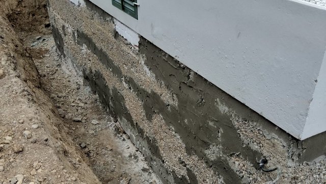 Freigelegte Hauswand mit grauer Masse und Kies im Erdgraben