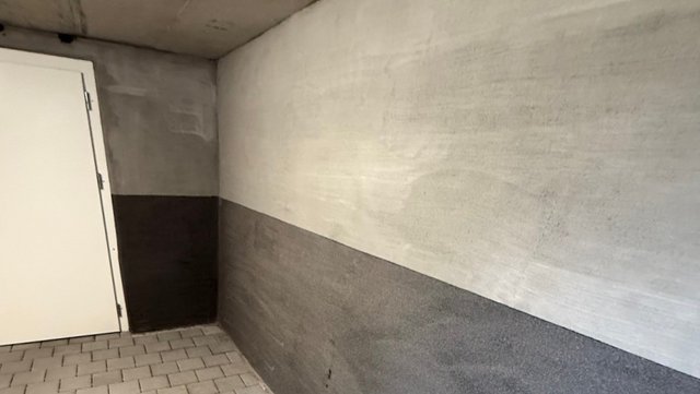 Weiß-grau gestrichene Kellerwand mit dunklem Sockelbereich und Tür, Fliesenboden