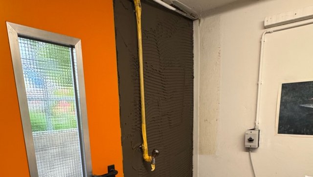 Graue Masse mit Zahnspachtel-Rillen auf Wand aufgetragen, gelbes Rohr verläuft senkrecht darüber.