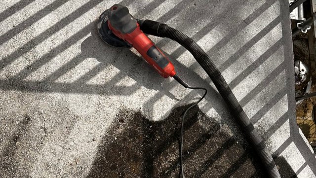 Rote Schleifmaschine mit Schlauch auf rauer Betonfläche, Schatten eines Geländers