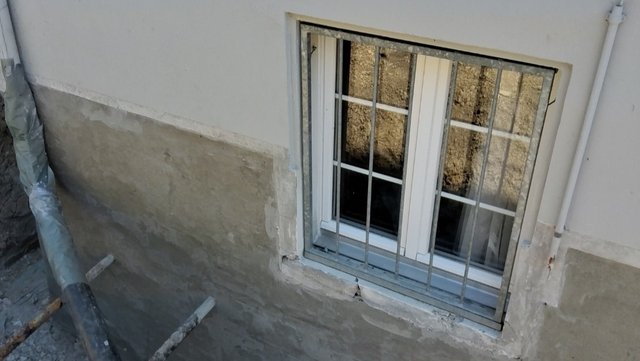 Kellerfenster mit Metallgitter, graue Masse an Wand und Fensterlaibung