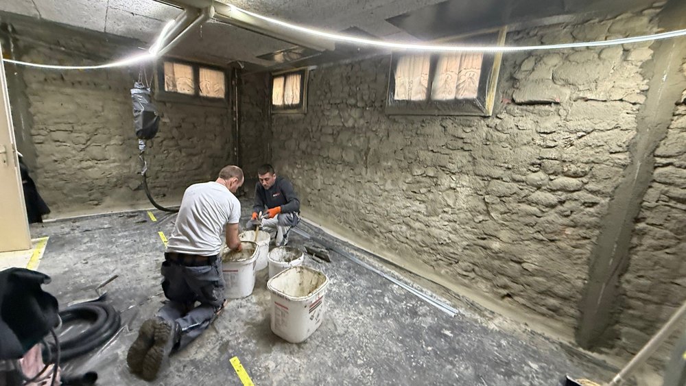 Zwei Arbeiter mischen graue Masse in Eimern vor freigelegter Kellersteinwand.