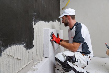 Arbeiter setzt helle Blöcke auf wandseitige graue Klebemasse.