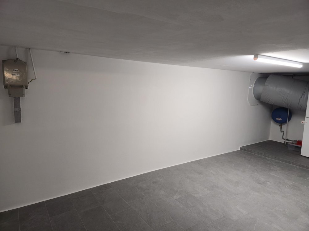 Heller Kellerraum mit weißer Wand, Fliesenboden, Lüftungsrohr und Leuchtstofflampe