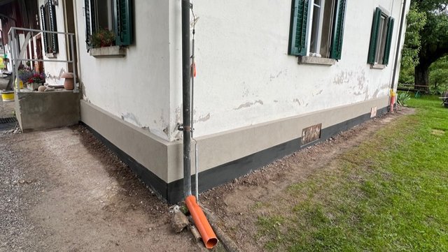 Freigelegte Sockelzone an Gebäudeecke mit grauer Beschichtung und dunklem Streifen, Drainagerohr am Fallrohr