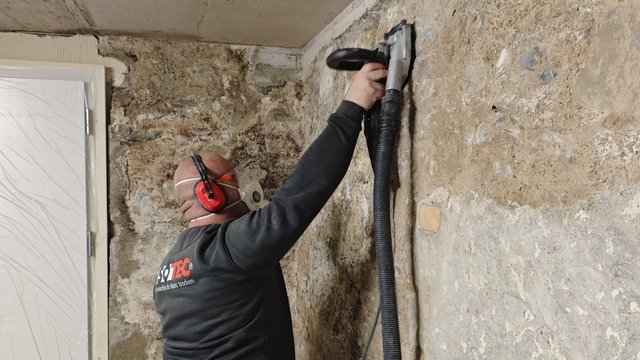 ISOTEC Mitarbeiter schleift Betonwand mit Handschleifer und Absaugschlauch