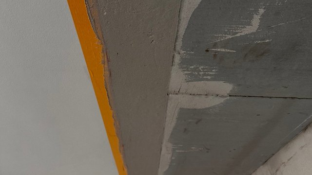 Nahaufnahme grauer Masse an Deckenkante, gelbes Abklebeband am Rand