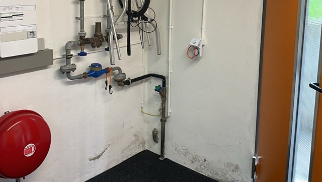 Technikraum mit Rohrleitungen, rotem Ausdehnungsgefäß und dunklen Feuchtflecken am Wandsockel