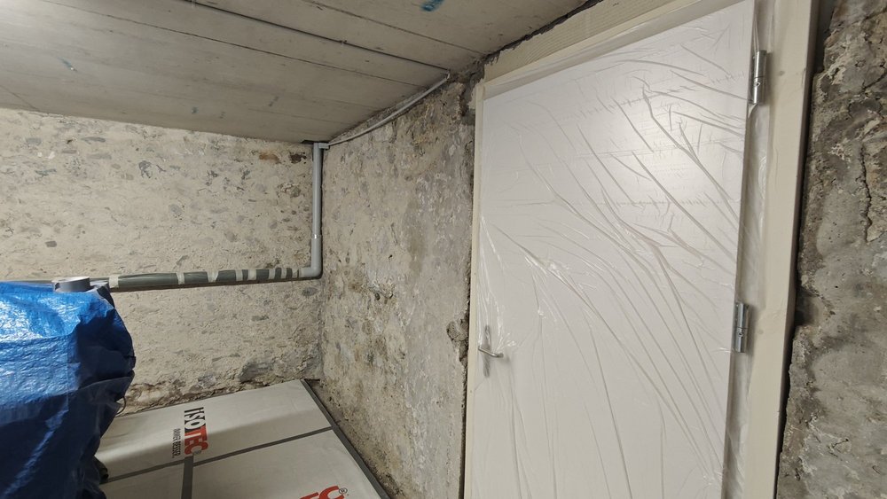 Kellerraum mit rauer Steinwand, Rohrleitung unter Decke und weißer Tür mit Schutzfolie