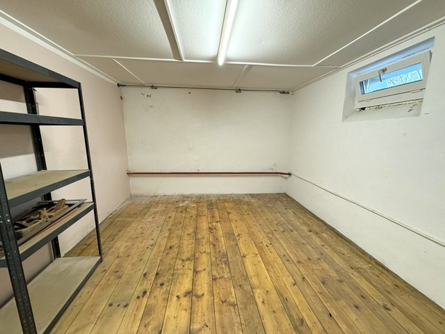 Leerer Kellerraum mit Holzbohlenboden, Regal links und kleinem Fenster mit Flecken am Rahmen