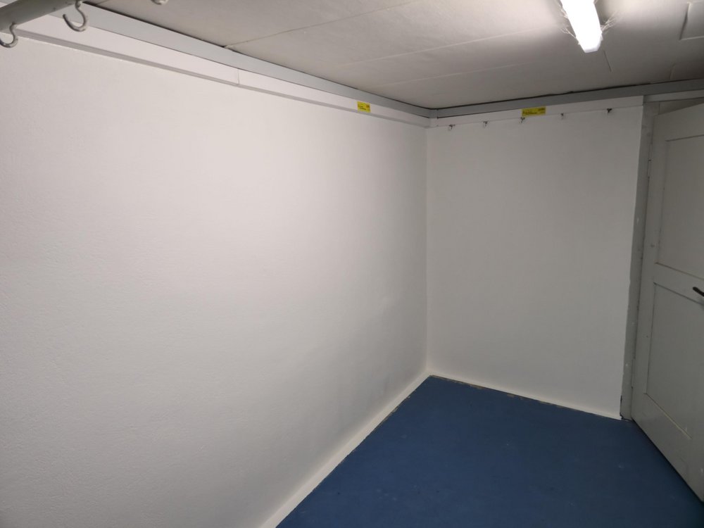 Weiß gestrichene Raumecke mit Deckenleuchte, grauer Tür und blauem Boden