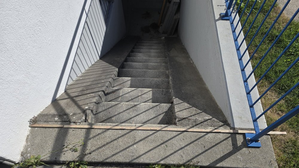 Betontreppe im Abgang, Zollstock quer auf dem Podest, blaues Geländer rechts.