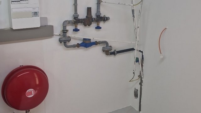 Technikraum mit rotem Ausdehnungsgefäß, grauen Rohrleitungen und blauem Wasserzähler an weißer Wand.