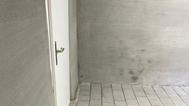 Graue Kellerwand mit dunklen Flecken neben weißer Tür, Fliesenboden im Vordergrund