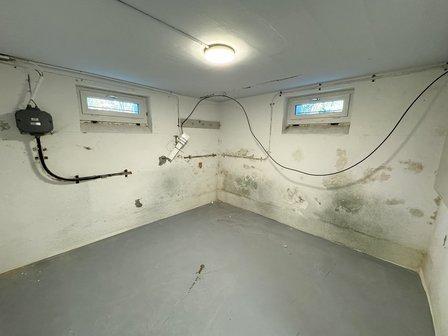 Kellerraum mit Flecken an Wand, zwei kleinen Fenstern und sichtbaren Kabeln.